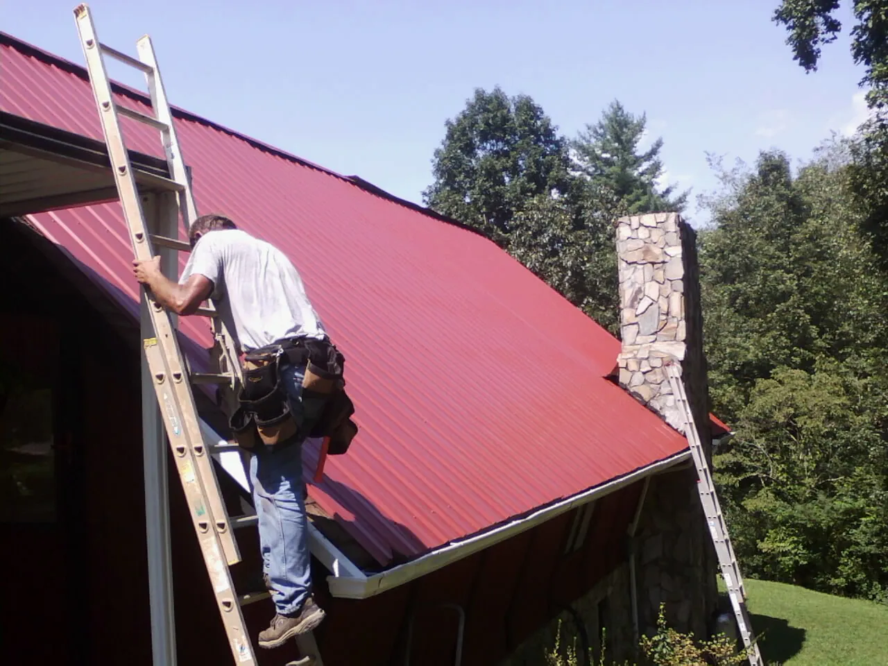 Roofer on a ladder beside a red metal roof and stone chimney — Soffit & Fascia Repair by Roofing Brothers in Jersey Shore